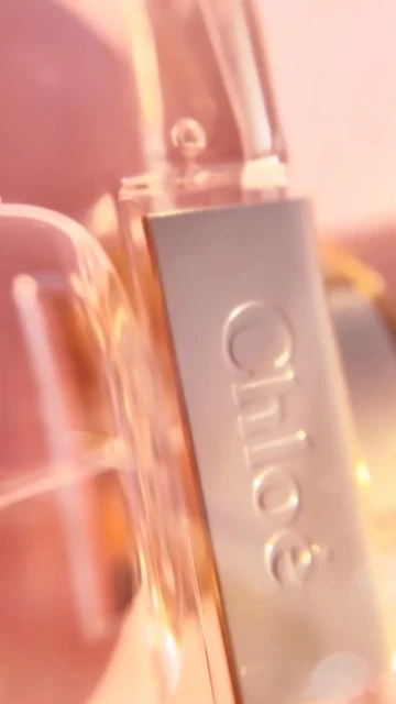 Close-up product shot of a bottle of Chloe perfume with a dropper, against a soft, diffused, pink-toned background and bright, diffused lighting.