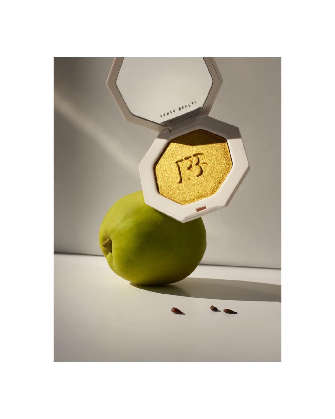 Fenty Beauty highlighter compact rests on a green pear with scattered maroon seeds, shot in studio with diffused light.