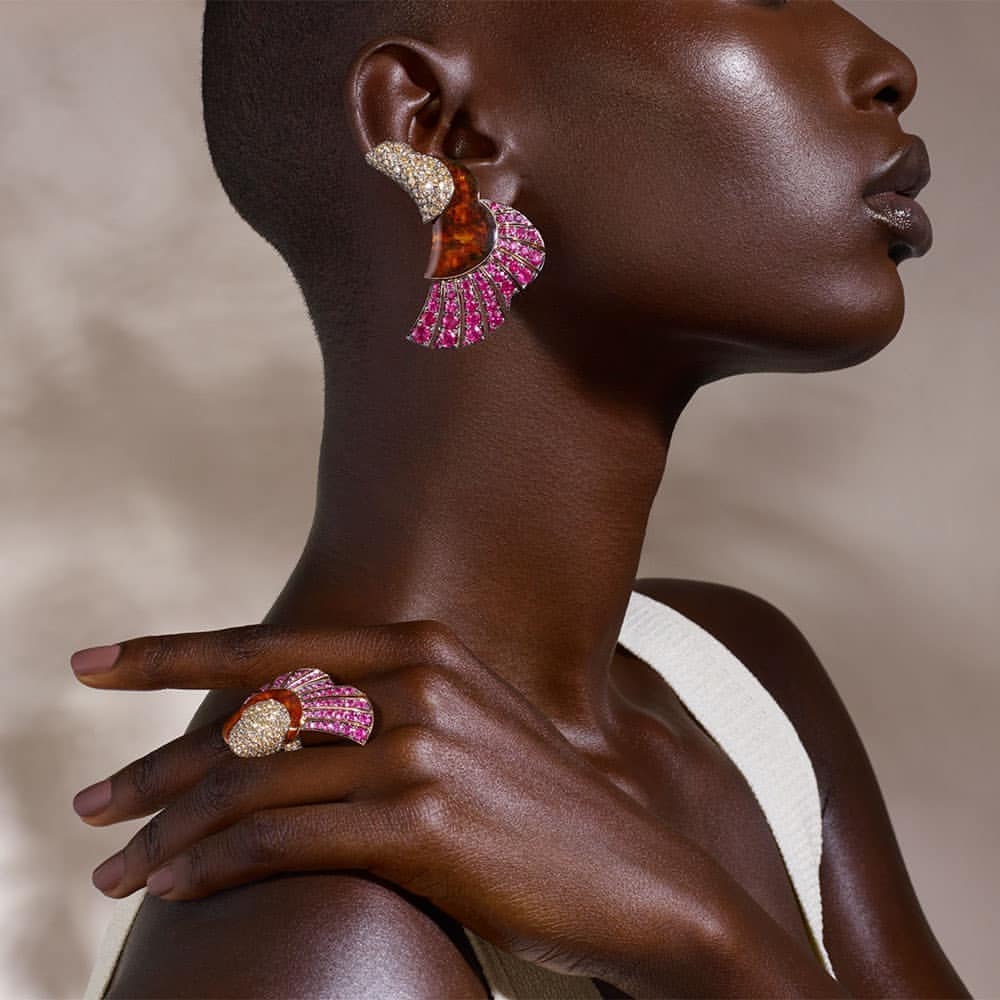 Close-up shot of a black woman modeling statement jewelry; a large ring and earring with gold, brown, and pink gemstones.