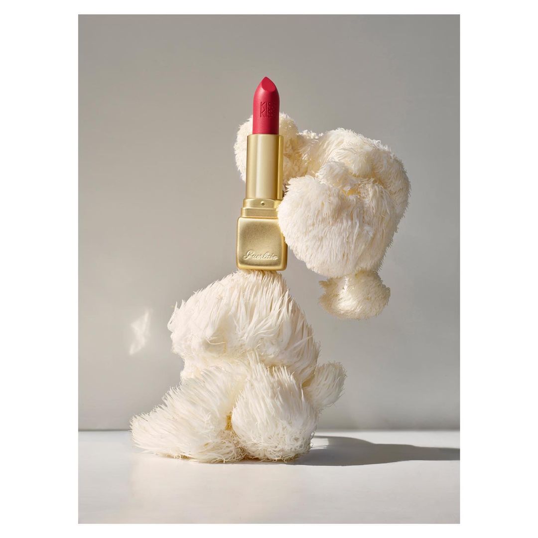 Elegant pink lipstick by Guerlain sitting on a fluffy white prop against a soft gray background in a beauty product shot.