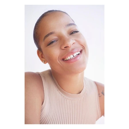 Close-up portrait of a smiling woman with a shaved head wearing a ribbed beige top against a white background