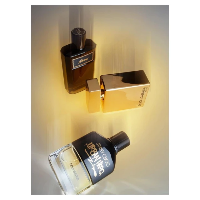 Three glass perfume bottles with black caps are displayed on a reflective surface in a studio, highlighted by soft, diffused lighting.