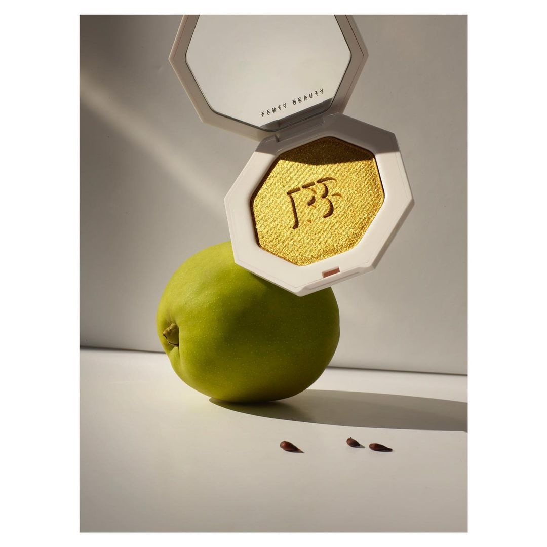 Fenty Beauty golden highlighter, lime green pear, and three small brown seeds on a white surface in a beauty product still life. 