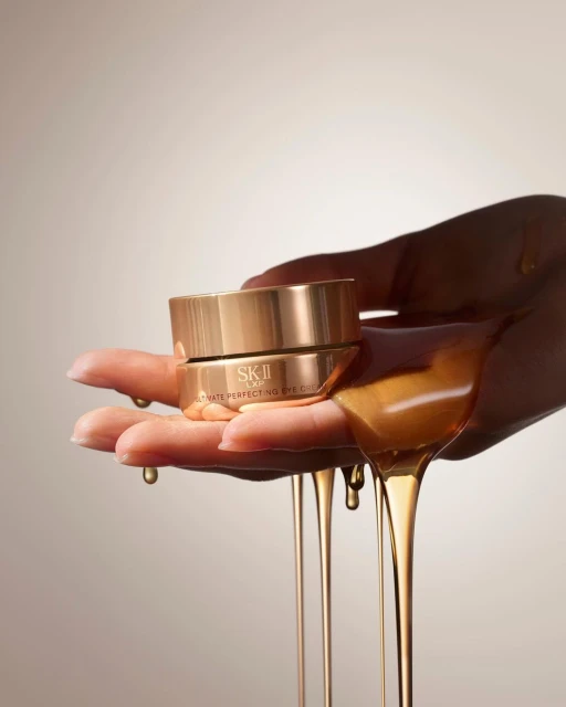 Close-up of a skincare product, SK-II Ultimate Perfecting Eye Cream, held in a hand with a honey-like substance dripping down against a tan background.