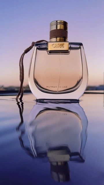 Close-up product shot of Chloé perfume bottle with gold accents and a reflection on a reflective surface in front of a purple sky.