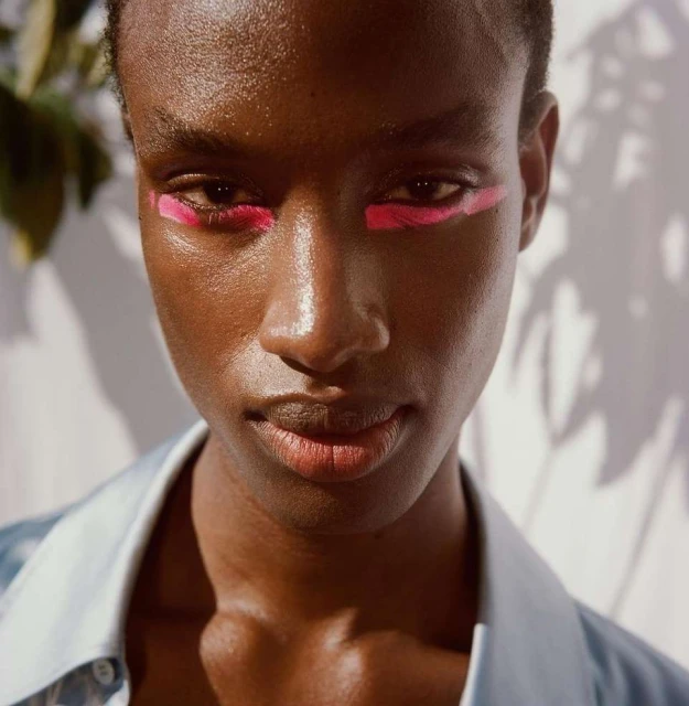 Headshot of a dark-skinned adult woman with bright pink graphic makeup, wearing a light blue shirt, and standing against a textured white background.