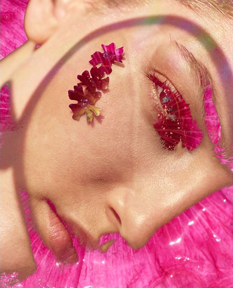 Close-up portrait of a fair-skinned adult female's face, adorned with flowers and artistic makeup, against a shimmering pink background.