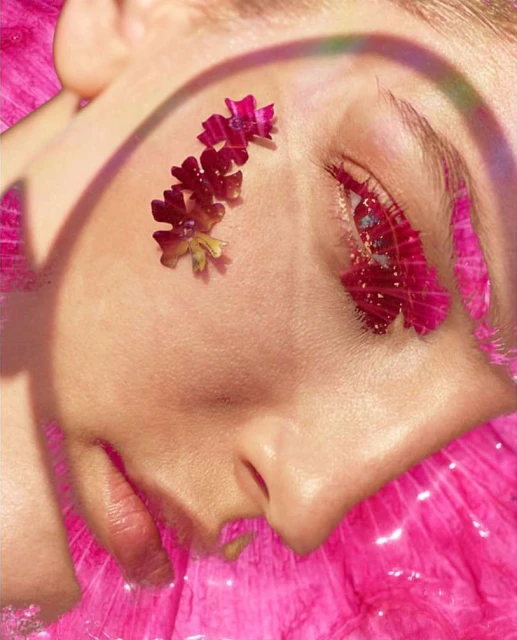 Close-up portrait of a fair-skinned adult female's face, adorned with flowers and artistic makeup, against a shimmering pink background.