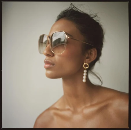 Portrait of a woman wearing oversized tinted sunglasses and pearl drop earrings against a plain light wall.