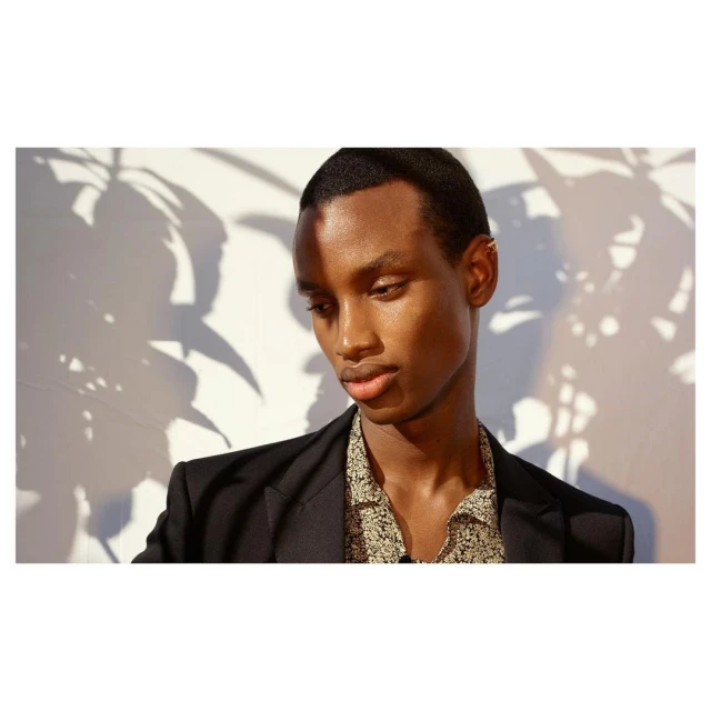 Close-up portrait of an African American adult male in a black blazer and patterned shirt, standing indoors in front of a white wall.