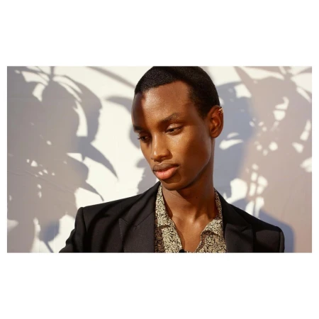 Close-up portrait of an African American adult male in a black blazer and patterned shirt, standing indoors in front of a white wall.