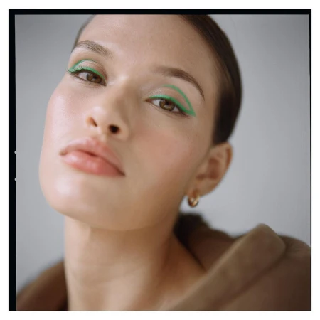 Close-up portrait of an adult female model with light skin and light brown hair, showcasing bright green eyeliner in a minimalist setting.