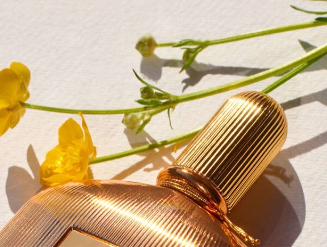 Gold perfume bottle with ribbed cap lies on a white surface next to a sprig of green leaves and bright yellow buttercup flowers.