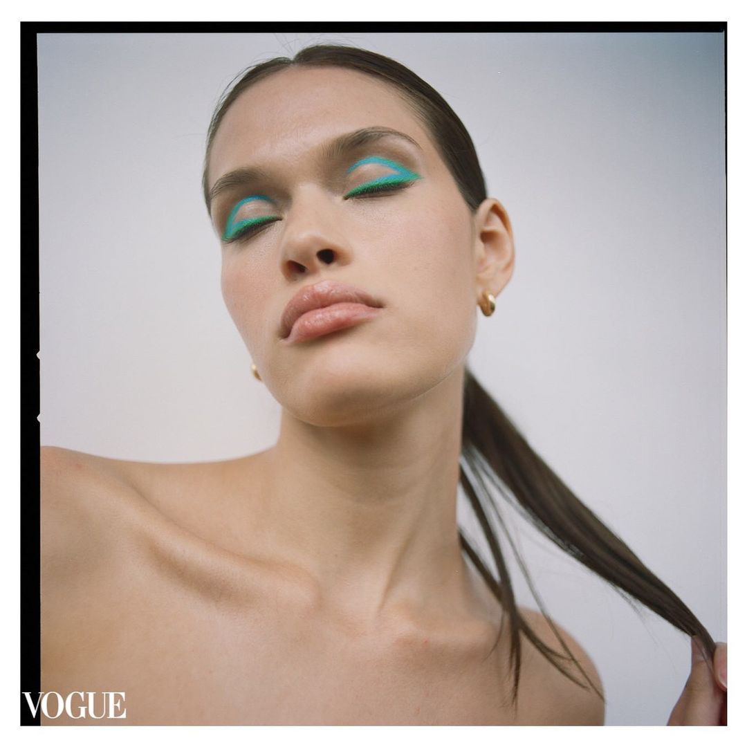 Close-up beauty shot of a model with teal eyeshadow and glossy lips against a neutral background; Vogue logo is visible in the corner.