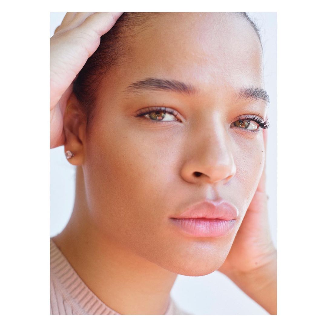 Close-up portrait of a light-skinned adult woman with brown eyes and hands near her face, wearing a light-colored top against a white background.