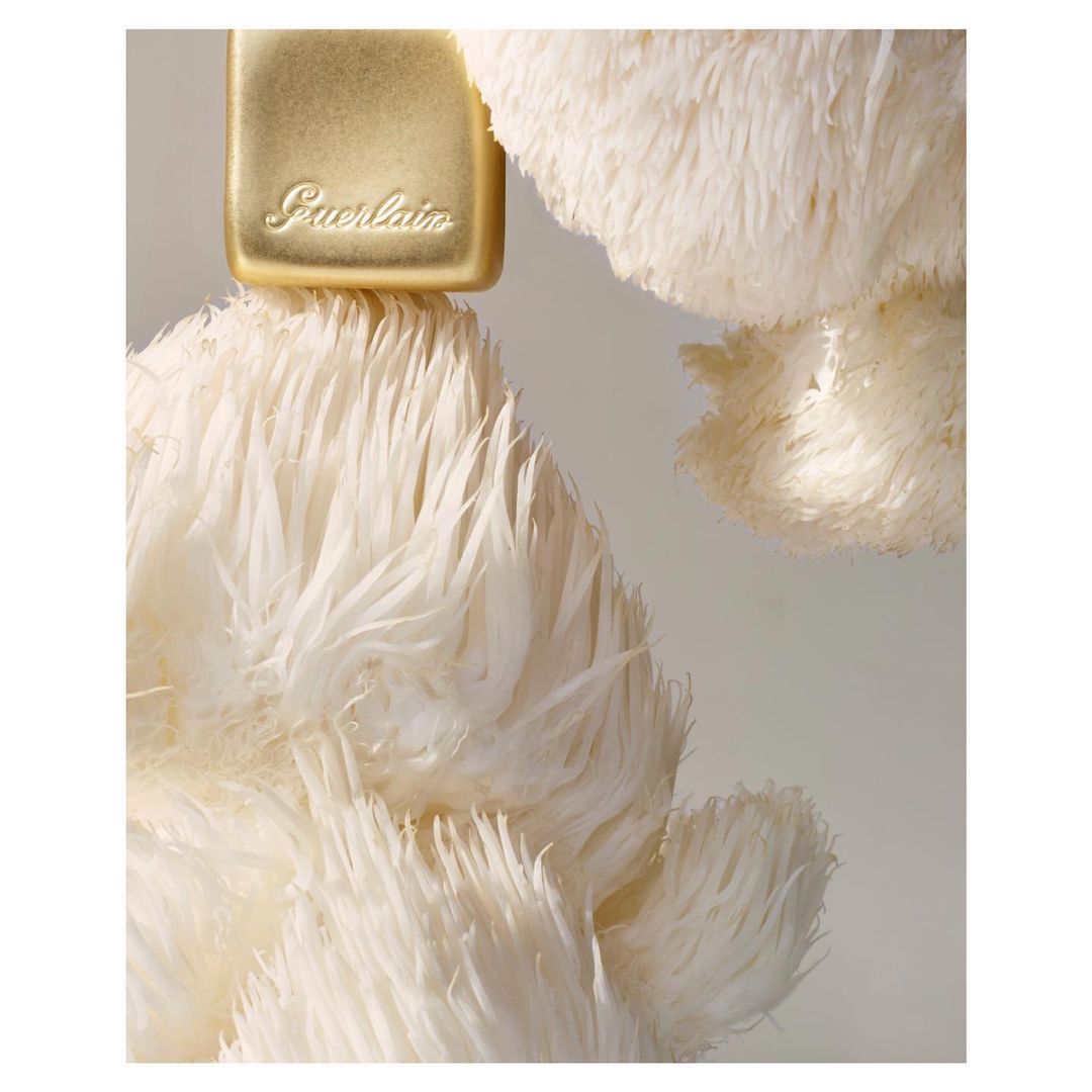 Close-up of a golden perfume bottle with the Guerlain logo, topped with fluffy white feathers, creating a soft and luxurious aesthetic.