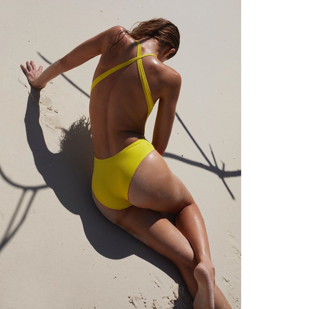 Woman in a yellow swimsuit is lying on a sandy beach on a sunny day. The swimsuit has an open back and crisscross straps.