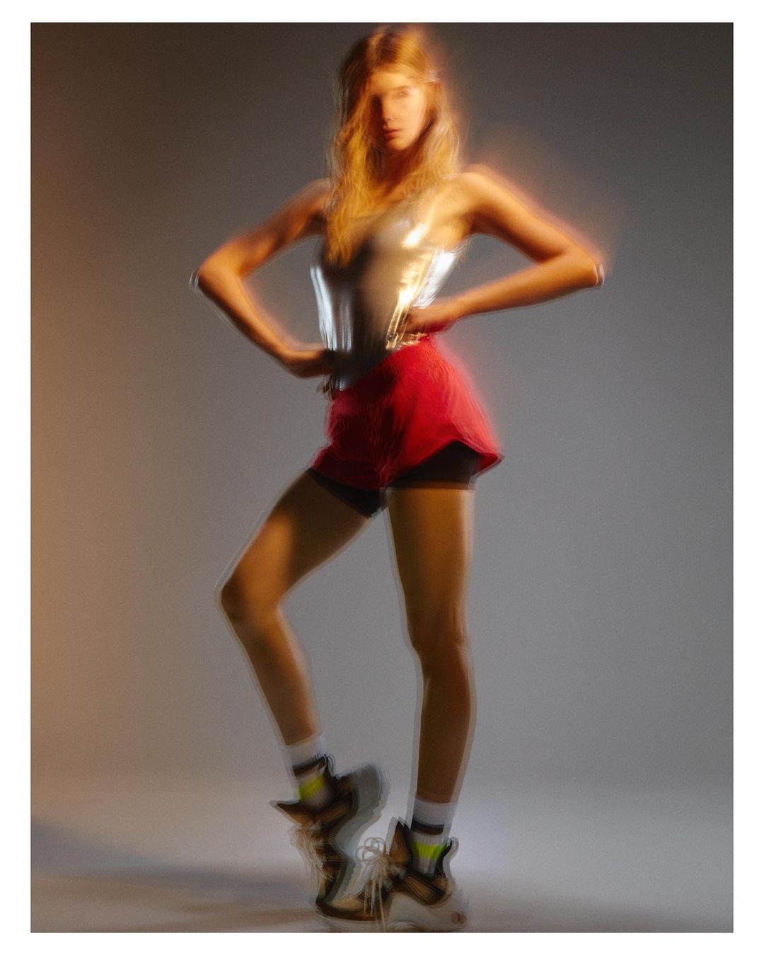 Model in silver top and red shorts posing against a grey gradient studio background with motion blur effect.
