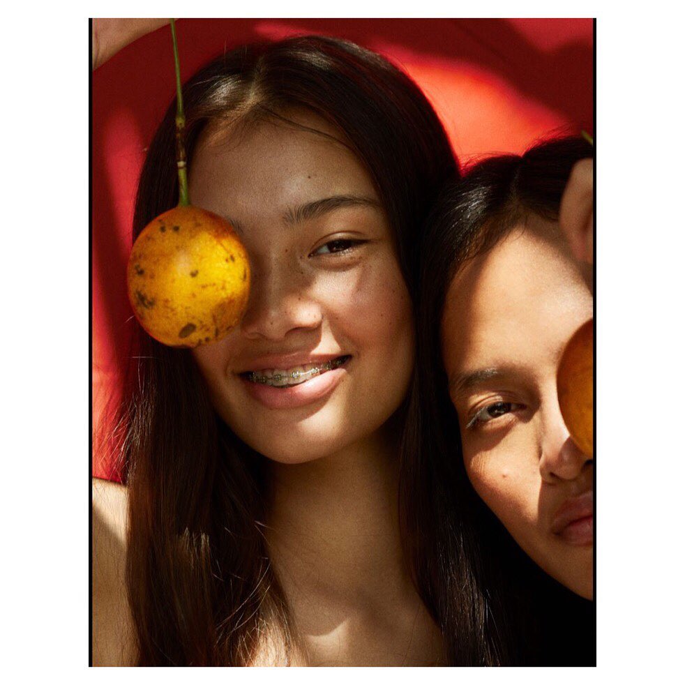 Two women pose intimately, one with braces, holding oranges against a vibrant red backdrop, bathed in warm, inviting studio lighting.