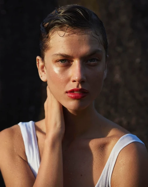 Close-up portrait of a young woman with wet slicked-back hair and red lipstick, posing against a dark rock formation backdrop.