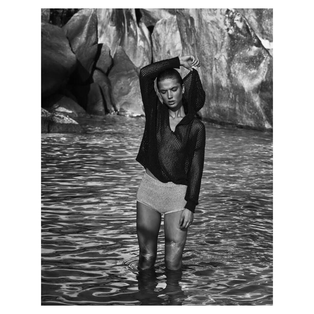 A young adult woman stands in the water wearing a mesh top and shorts, with large rocks and trees in the background, black and white.