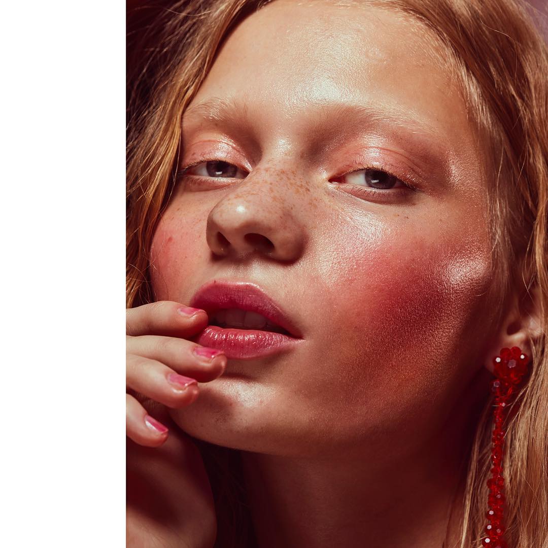 Close-up of a young woman with red-toned makeup posing indoors, showcasing pink eyeshadow, bright blush, and red dangling earrings.