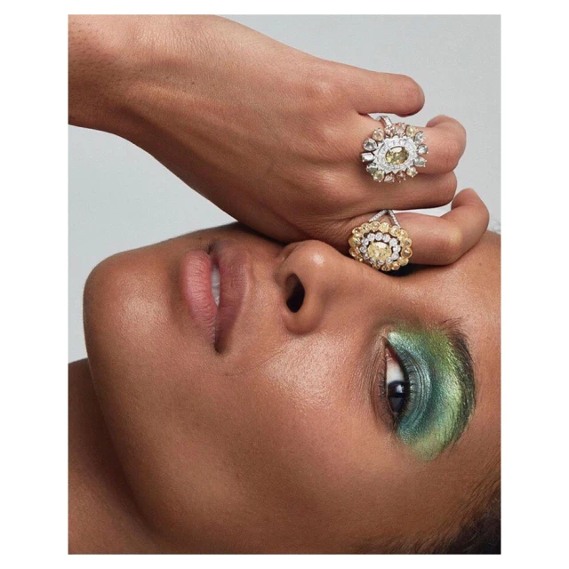 Close-up portrait of a woman with tan skin, green eyeshadow, and two floral rings, set against a plain light gray background.
