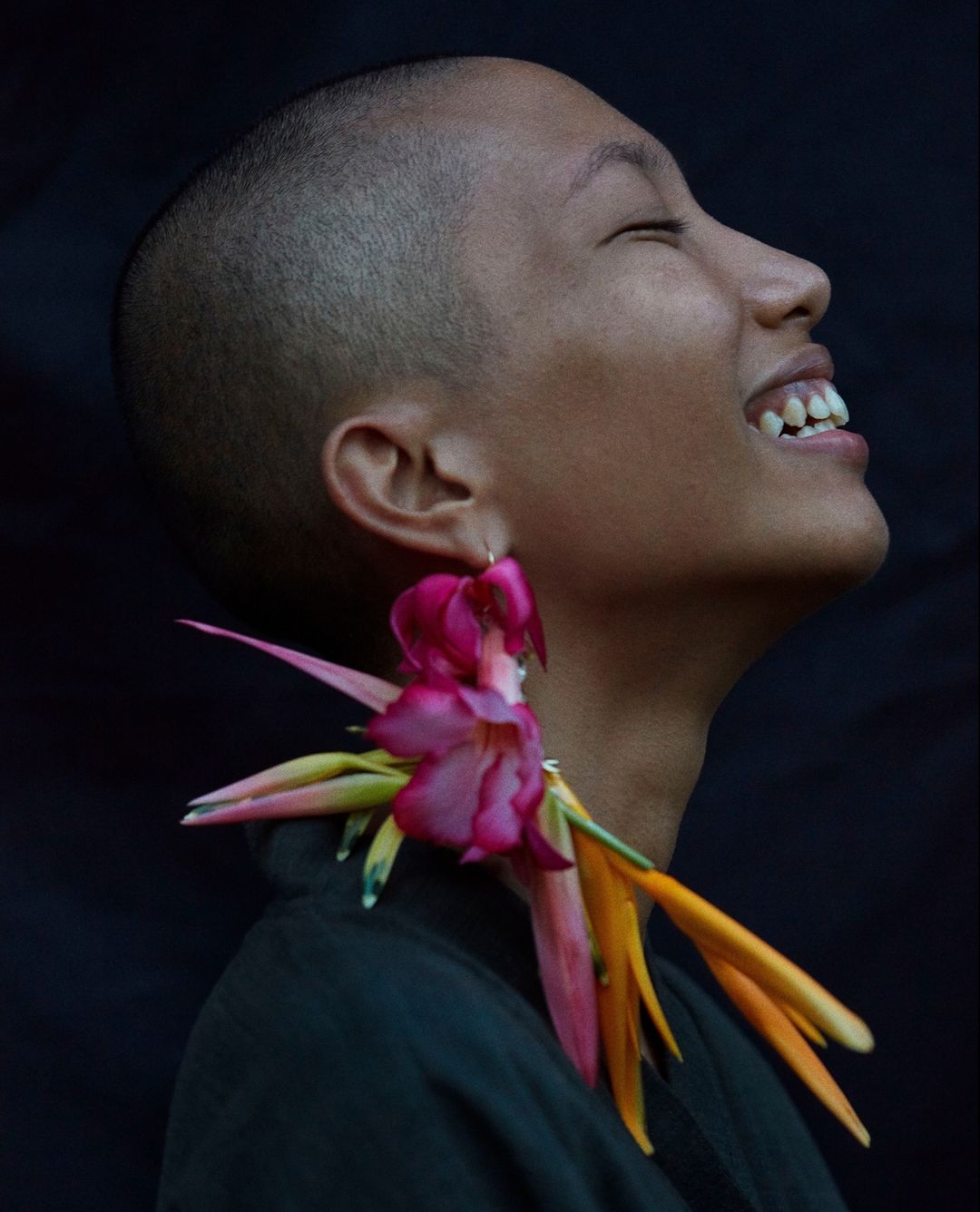 Close-up profile shot of a smiling woman with short hair, large floral earrings, wearing a dark shirt against a dark background.