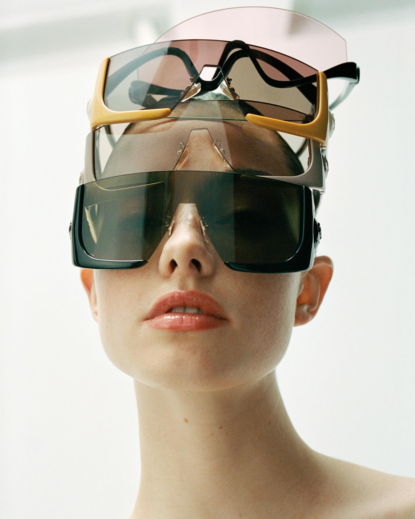 Close-up of a young woman with fair skin wearing a stack of four sunglasses of various styles, against a plain white backdrop.