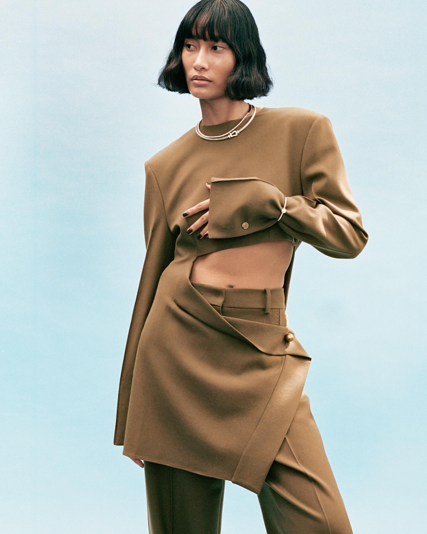 Fashion-forward portrait of an Asian female model in a stylish brown cut-out suit against a light blue backdrop in a photo studio.