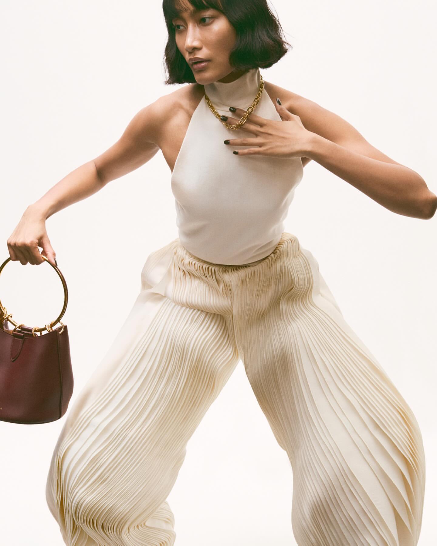 Adult female fashion model in a light halter top and pleated pants holding a bucket bag in a studio environment on a white background.
