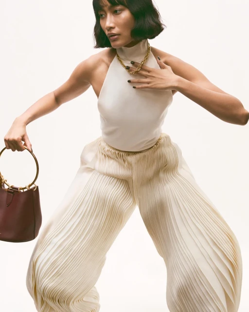 Adult female fashion model in a light halter top and pleated pants holding a bucket bag in a studio environment on a white background.
