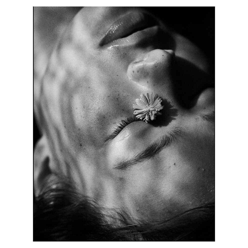 Close-up black and white portrait of a person with a daisy on their eyelid, with light patterns across the face and hair visible.