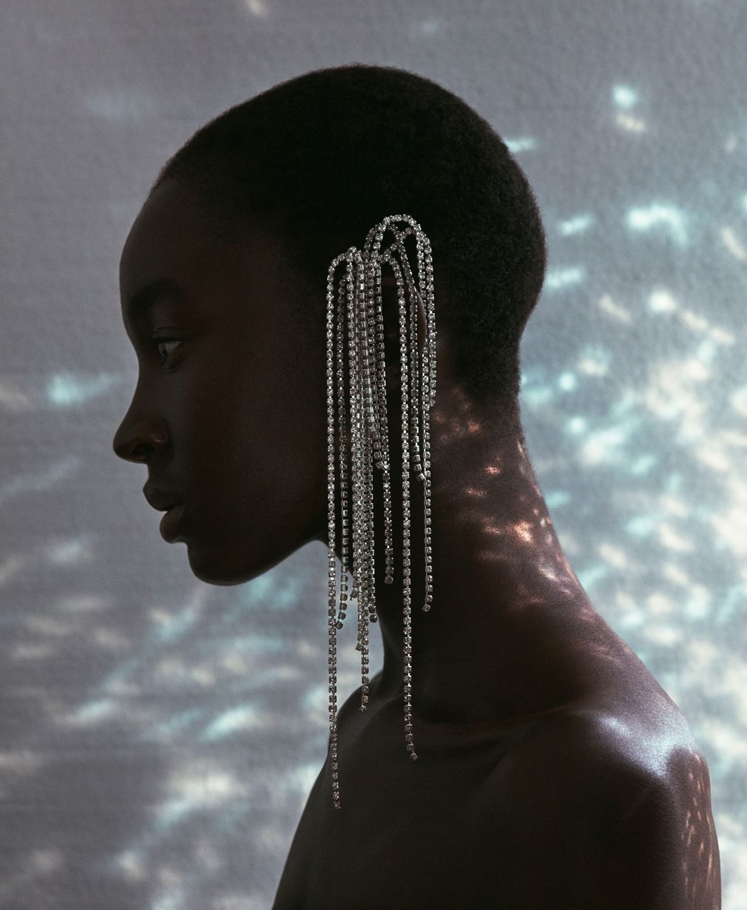 Profile portrait of a Black woman with short hair wearing an elaborate crystal fringe ear cuff against a wall with dappled light patterns.