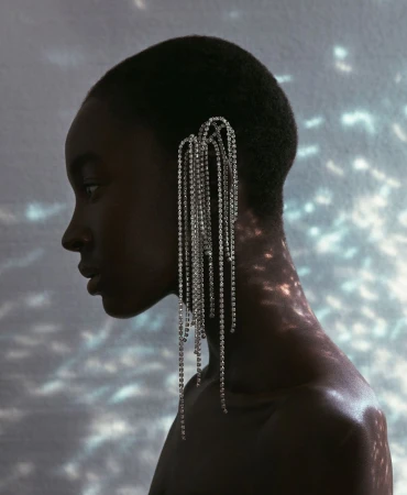 Profile portrait of a Black woman with short hair wearing an elaborate crystal fringe ear cuff against a wall with dappled light patterns.