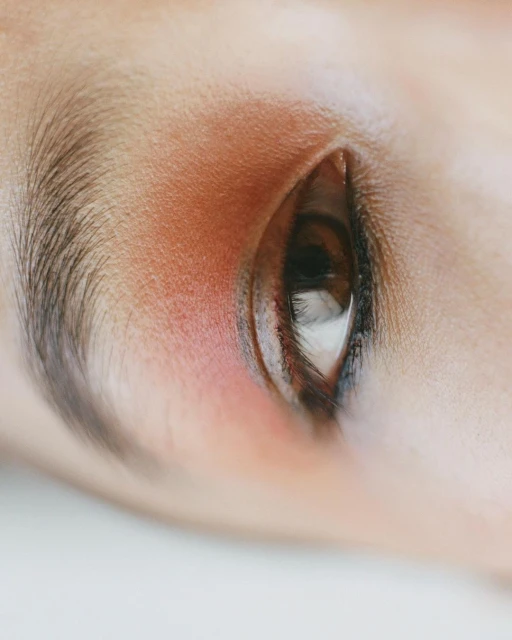 Macro close-up of a woman's eye with red eyeshadow. The image focuses on the eye, lashes, and surrounding skin.