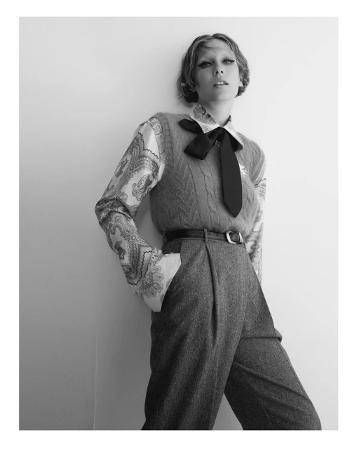 Black and white portrait of a young adult woman in a patterned shirt, sweater vest, wide-legged trousers, and tie posing indoors.