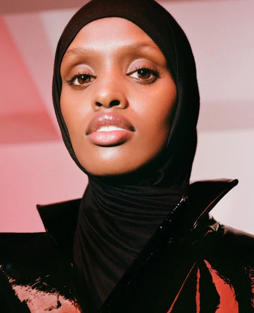 Close-up portrait of a woman with dark skin, wearing a black hijab and shiny black jacket, posing against a gradient background of white and pink.