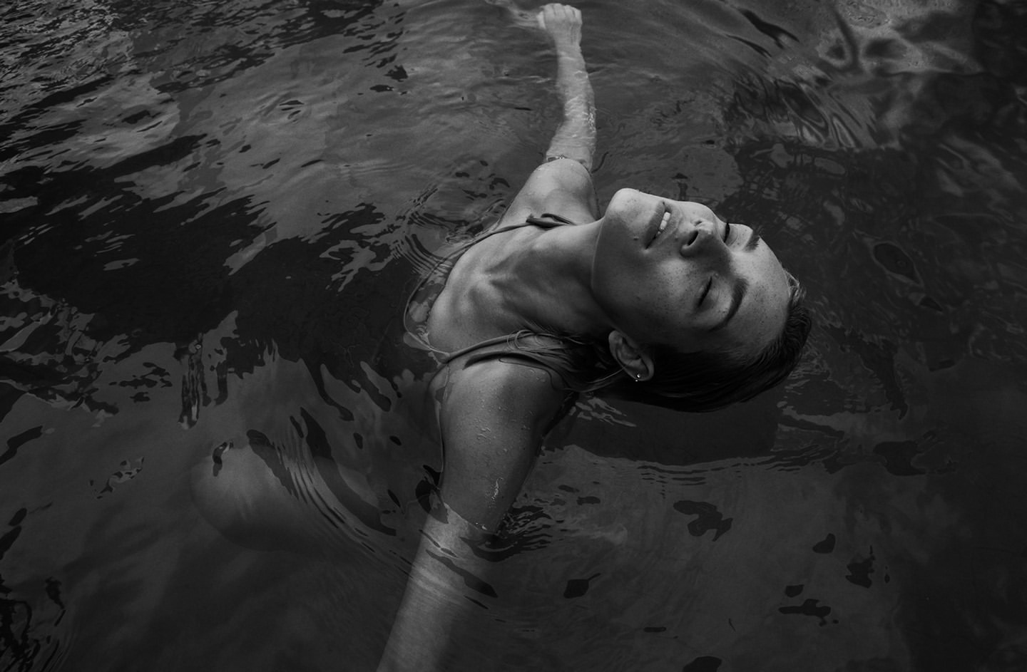 Monochrome portrait of a woman floating in water with eyes closed, creating a serene and tranquil atmosphere in an outdoor setting.