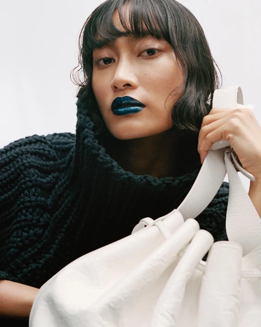 Close-up of an Asian woman with dark curly hair and bold blue lipstick wearing a black knit sweater and holding a white leather bag.