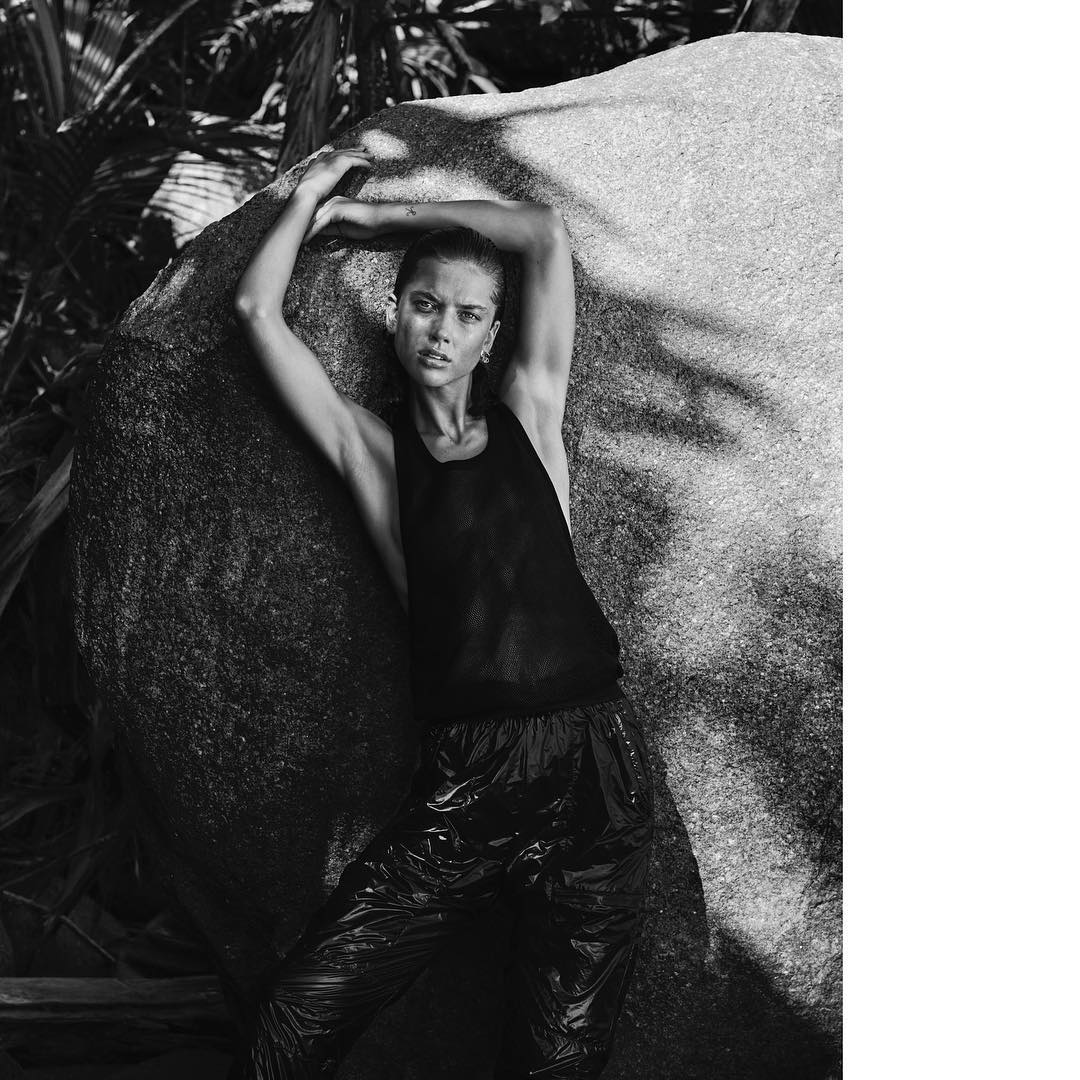 Black-and-white portrait of a woman posing near a large rock formation in a tropical environment, wearing a tank top and shiny pants.
