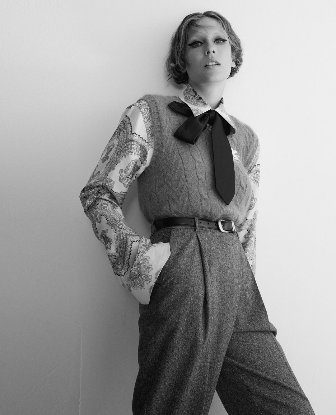 Monochrome fashion shot of a female model with a black bow tie, wearing wool slacks and vest leaning against a white wall.