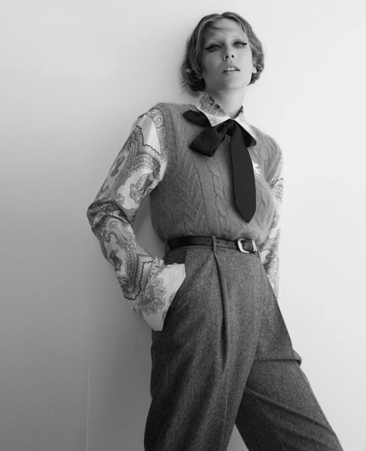 Monochrome fashion shot of a female model with a black bow tie, wearing wool slacks and vest leaning against a white wall.