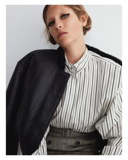 Woman in striped button-down shirt and black jacket posing against a white background in a medium shot with soft studio lighting.