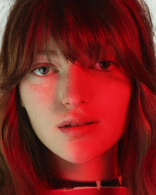 Close-up portrait of a young woman with bangs, lit by a vibrant red light, highlighting her face and hair against a bright background.