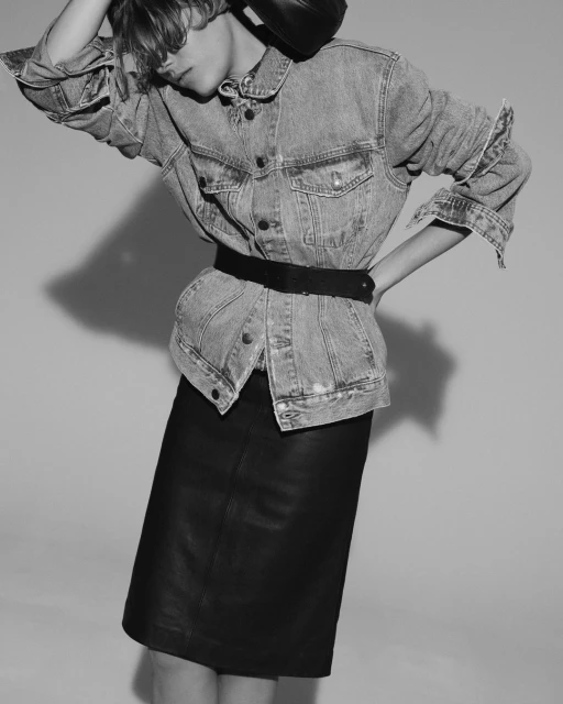 A woman in a denim jacket and black skirt strikes a pose. The black and white image features a fashion-forward aesthetic and studio lighting.