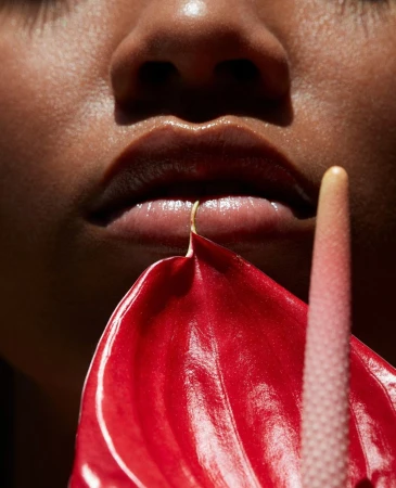 Close-up of a woman's face with shiny skin, red anthurium flower near lips, and a dark background. Focus on beauty and skincare.