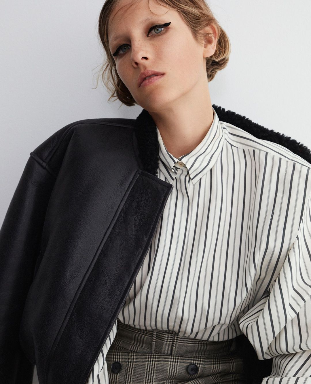 Woman in a striped shirt and black jacket posing indoors, showcasing fashion and beauty with a neutral background and soft lighting.