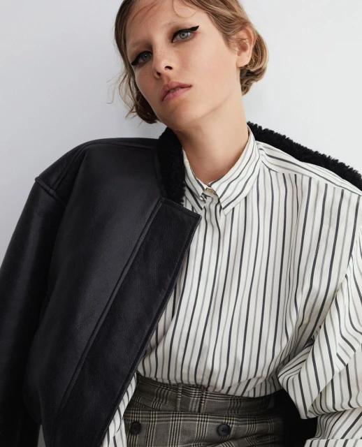 Woman in a striped shirt and black jacket posing indoors, showcasing fashion and beauty with a neutral background and soft lighting.
