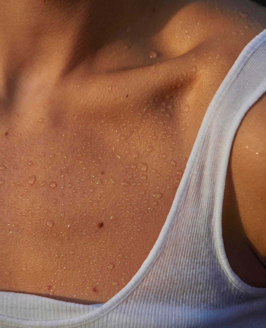 Close-up of a fair-skinned adult female's upper chest and shoulders glistening with water droplets, wearing a white tank top.